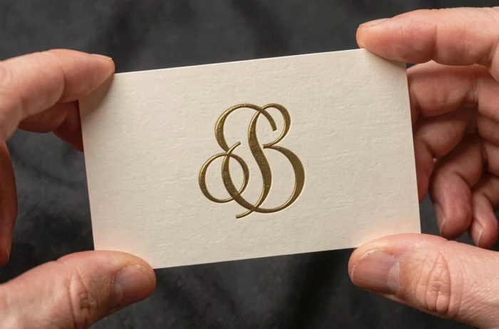 Carte de visite embossée tenue entre deux doigts affichant un monogramme doré formé de deux lettres entrelacées